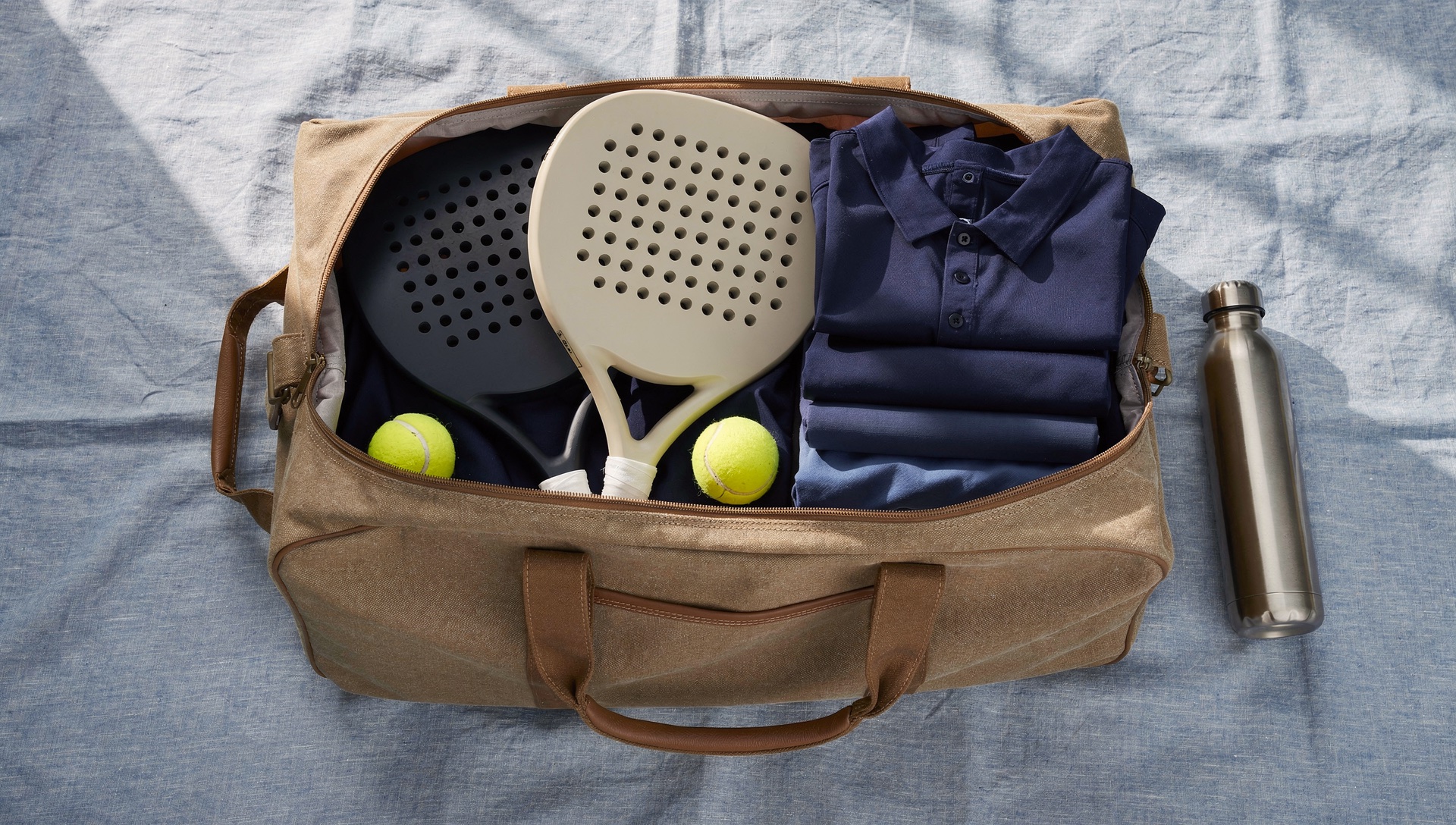 AI illustration of padel rackets, yellow balls and folded navy clothes packed in a tan travel bag.